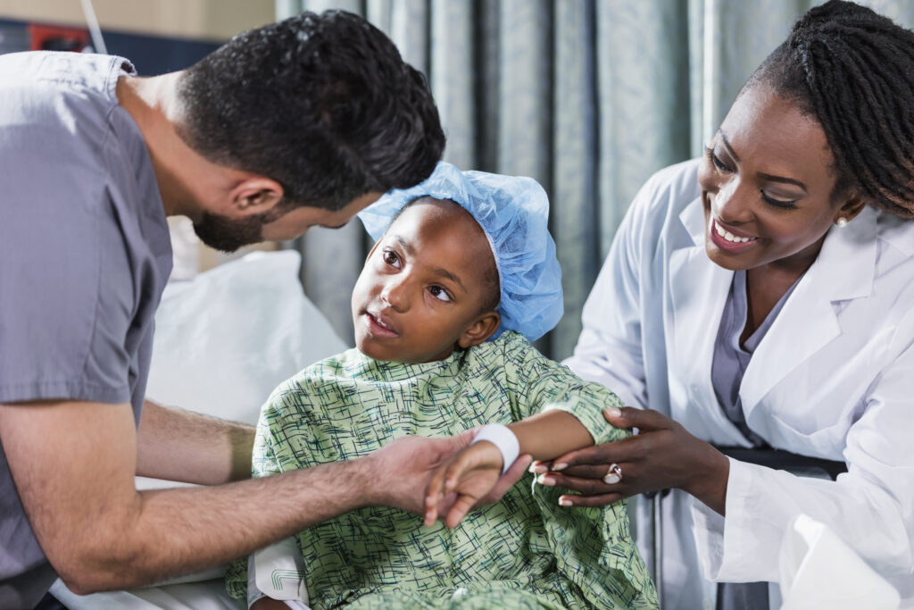 A 6 year old African-American girl sitting on a hospital gurney, with a doctor and nurse tending to her. She is wearing a hospital gown and surgical cap. A nurse in scrubs, a young Middle Eastern man in his 20s, is standing on one side of her, and a female doctor, a mature African-American woman in her 40s, is standing on the other side.